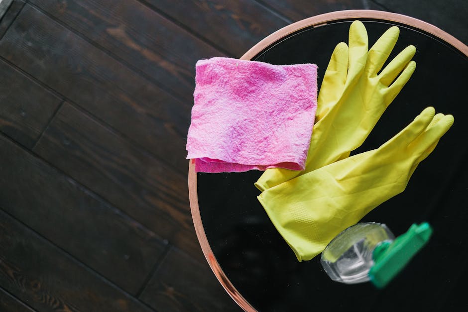 A round black table with a wooden edge, positioned on dark wooden flooring, holds a pink microfiber cloth, a pair of yellow rubber gloves, and a clear spray bottle with a green nozzle. The scene depic