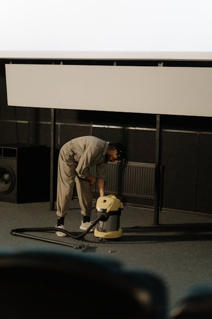 Close-up of a vacuum cleaner with a transparent dust container filled with debris, connected to a flexible, black and white patterned hose. The vacuum is positioned on a pinkish-beige carpet in a room